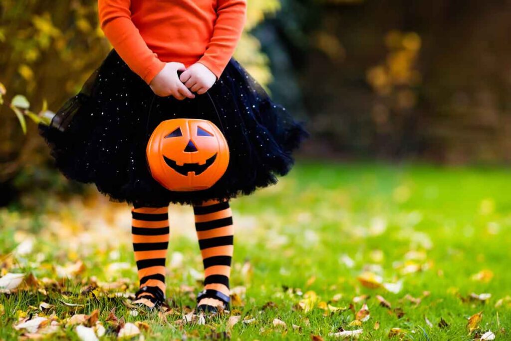 Small child wearing a black sparkly tutu, orange long sleeve shirt, black and orange striped tights, and black Mary Janes. They are holding a pumpkin Trick-or-Treat bucket and standing in grass with dead leaves all around.