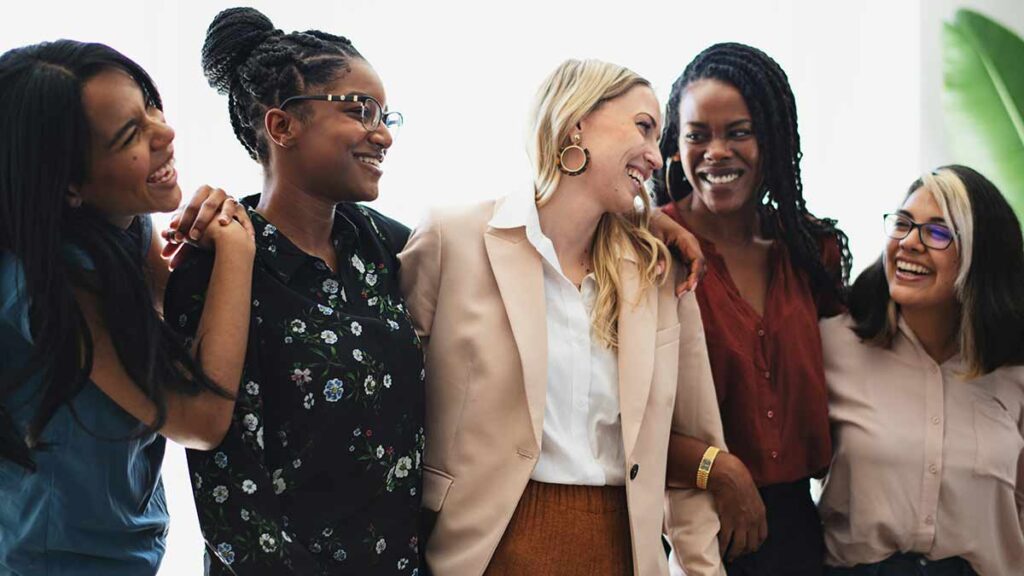 Five businesswomen stand with arms around each other.