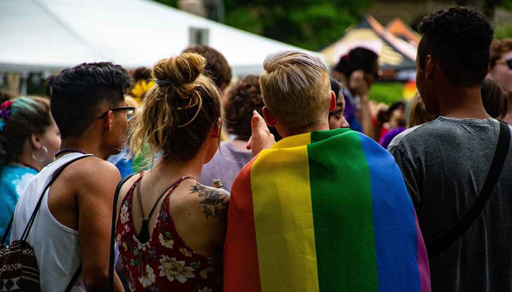 A group of friends in a crowd look at the festivities around them. A young male wears the Pride flag around his shoulders.